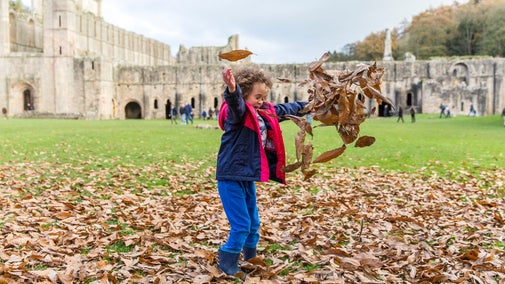 Child playing in the fallen leaves in the park at Fountains Abbey and Studley Royal Water Garden, North Yorkshire